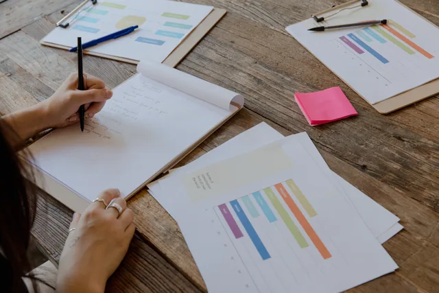 Hands writing on paper with colorful charts on wooden table, organized workspace. Customer data platforms selection checklist: entity and edition